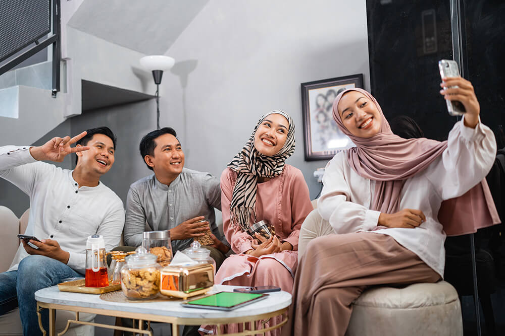 A group of four smiling friends, two men and two women, taking a selfie in a modern living room. On the coffee table in front of them are traditional snacks and drinks, indicating a Hari Raya celebration.