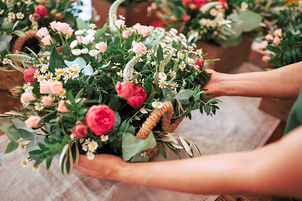 Hands arranging a floral bouquet with a mix of pink roses, daisies, and lush greenery on a table.